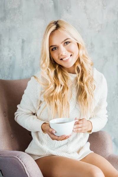 Усміхнена приваблива жінка в светрі сидить на кріслі і тримає чашку кави в спальні вранці — стокове фото
