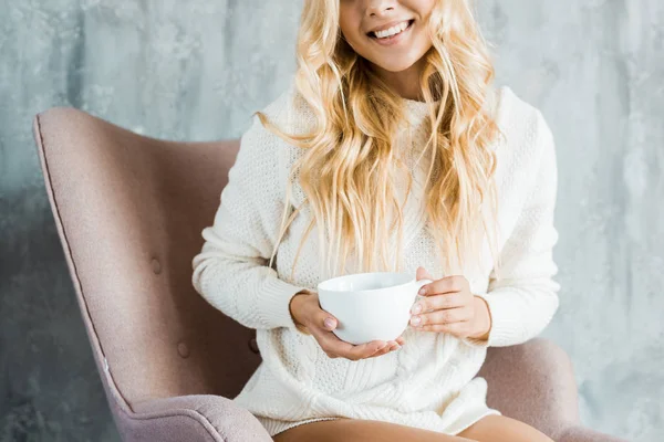 Imagem cortada de mulher em suéter sentado em poltrona e segurando xícara de café no quarto de manhã — Fotografia de Stock