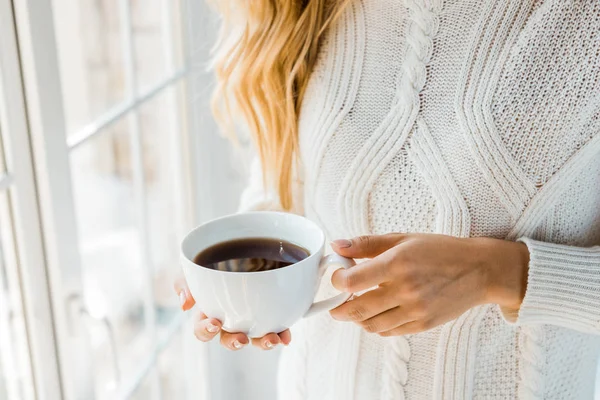 Обрезанное изображение женщины в свитере с чашкой кофе у окна дома — стоковое фото