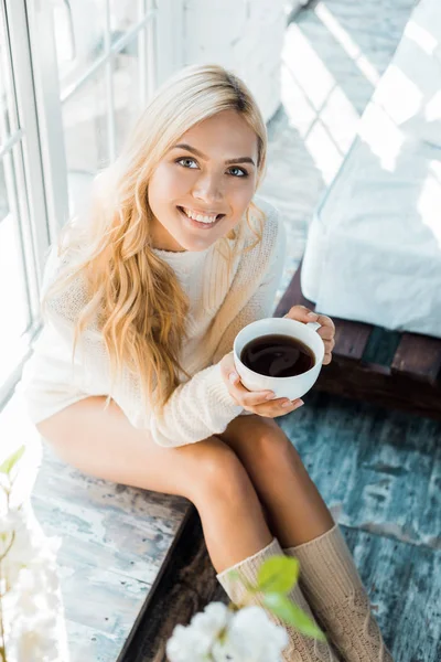 Visão de alto ângulo de mulher atraente sorridente em suéter segurando xícara de café no quarto de manhã — Fotografia de Stock