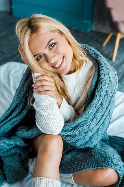 Улыбающаяся привлекательная женщина в свитере сидит завернутая в одеяло и смотрит на камеру в спальне утром — стоковое фото