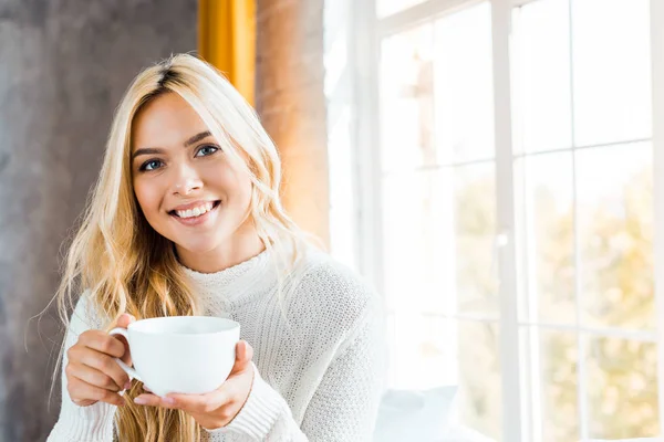 Fröhliche schöne Frau im Pullover hält Tasse Kaffee in der Hand und blickt morgens in die Kamera im Schlafzimmer — Stockfoto