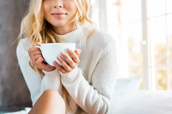 Imagen recortada de la mujer en suéter sosteniendo taza de café en el dormitorio por la mañana - foto de stock