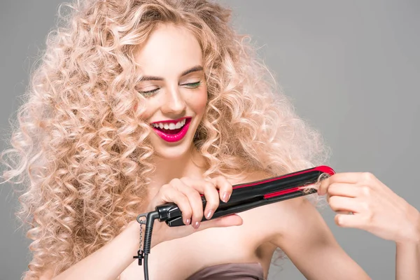 Smiling young woman with long curly hair holding hair straightener isolated on grey — Stock Photo