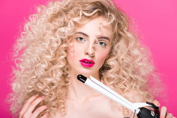 Bela jovem com cabelos longos encaracolados segurando encrespador de cabelo e olhando para a câmera isolada em rosa — Fotografia de Stock