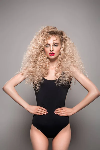 Beautiful young woman with long curly hair standing with hands on waist and looking at camera on grey — Stock Photo