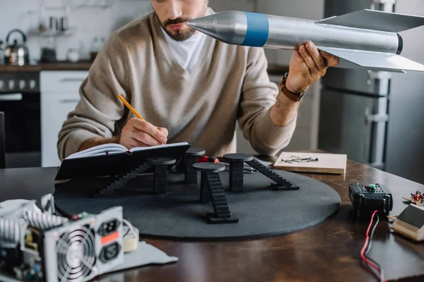 Imagen recortada del ingeniero sosteniendo el modelo de cohete y tomando notas en casa - foto de stock