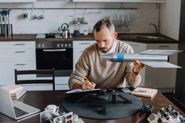 Engenheiro bonito segurando modelo de foguete e fazendo anotações em casa — Fotografia de Stock