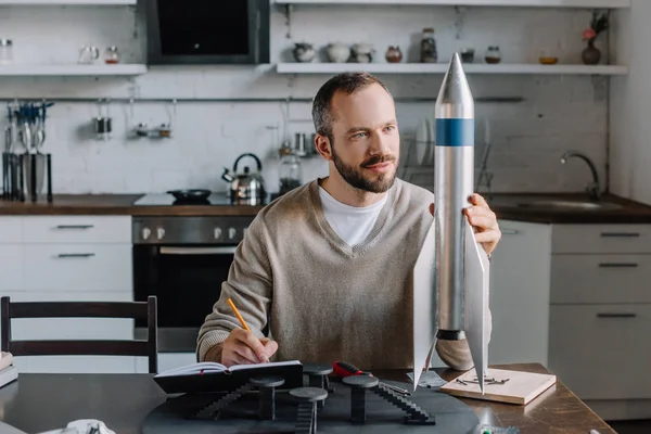 Schöner Ingenieur schaut sich Raketenmodell an und macht Notizen zu Hause — Stockfoto