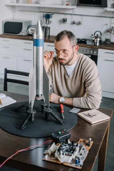 Schöner Ingenieur blickt über Brille auf Raketenmodell zu Hause — Stockfoto