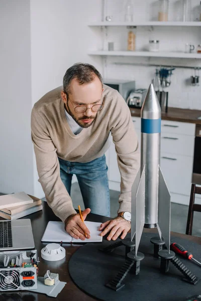 Handsome engineer looking at rocket model and making notes at home — Stock Photo