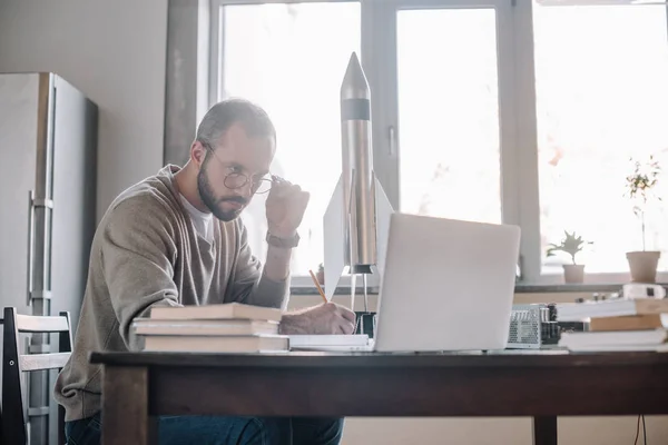 Schöner Ingenieur modelliert Rakete und schaut zu Hause auf Laptop — Stockfoto