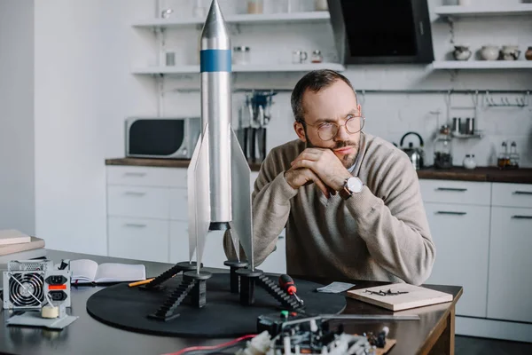 Thoughtful engineer sitting at table with rocket model at home and looking away — Stock Photo