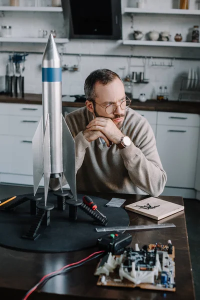 Nachdenklicher Ingenieur sitzt zu Hause mit Raketenmodell am Tisch und schaut weg — Stockfoto