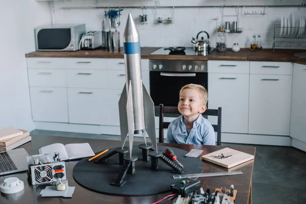 Fröhlicher liebenswerter Junge sitzt am Wochenende mit Raketenmodell in der Küche am Tisch — Stockfoto