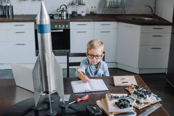 Adorable boy modeling rocket and writing something to notebook in kitchen on weekend — Stock Photo