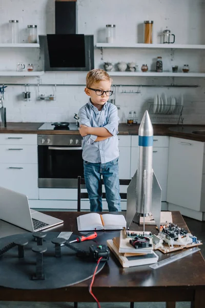 Adorable garçon debout avec les bras croisés et regardant modèle de fusée dans la cuisine le week-end — Photo de stock
