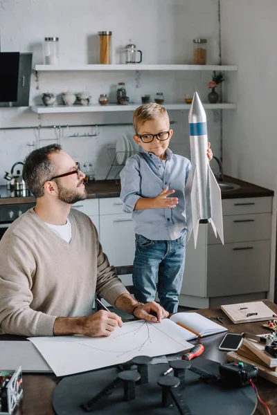 Lächelnder Sohn hält Raketenmodell zu Hause am Tisch — Stockfoto