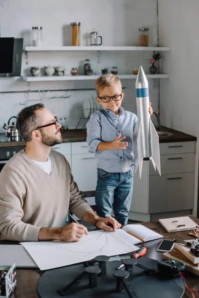 Sonriente hijo sosteniendo cohete modelo en la mesa en casa - foto de stock