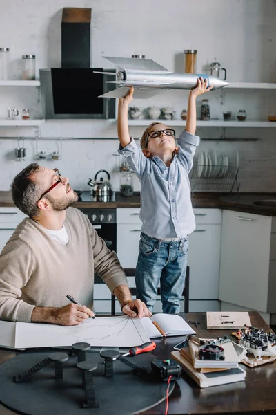 Alegre filho segurando modelo de foguete na mesa em casa — Fotografia de Stock