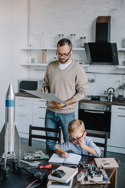 Nachdenklicher Vater schaut auf Blaupause und Sohn schreibt zu Hause etwas ins Notizbuch — Stockfoto