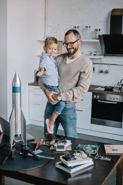 Sonriente padre llevando hijo y mirando el modelo de cohete en casa - foto de stock