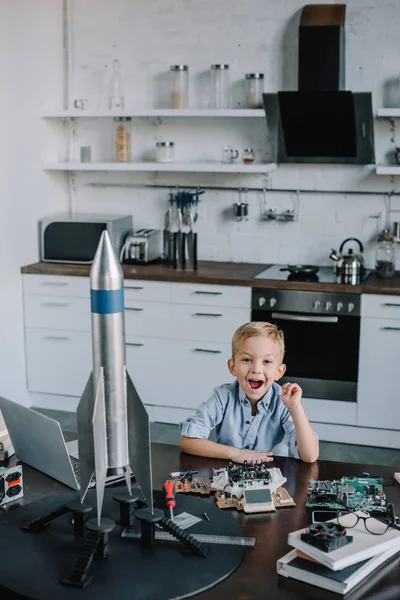 Happy adorable boy sitting at table with rocket model in kitchen and looking at camera — Stock Photo