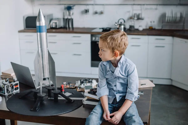Irritated grimacing boy sitting on table near rocket model in kitchen on weekend — Stock Photo