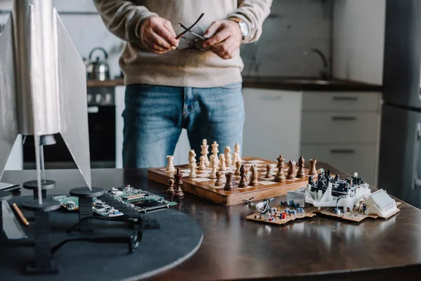 Image recadrée de l'homme debout près de la table avec un échiquier et tenant des lunettes à la maison — Photo de stock