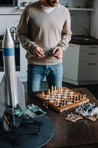 Imagen recortada del hombre de pie cerca de la mesa con tablero de ajedrez en casa - foto de stock