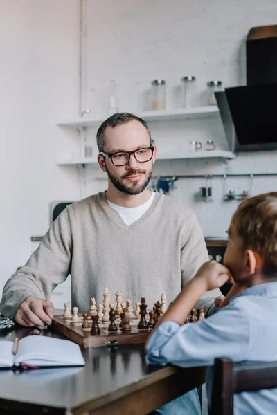 Батько в окулярах дивиться на маленького сина, граючи в шахи разом вдома — стокове фото