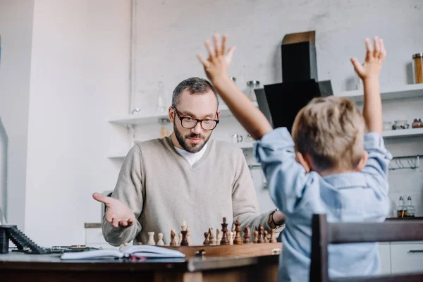 Hijo pequeño triunfante levantando las manos mientras juega al ajedrez con el padre en casa - foto de stock