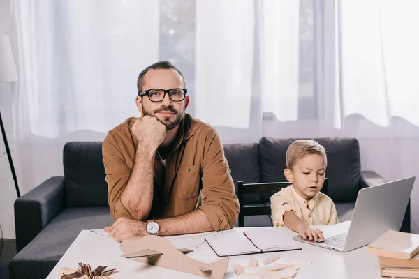 Thoughtful father in eyeglasses looking at camera and little son using laptop while modeling together at home — Stock Photo