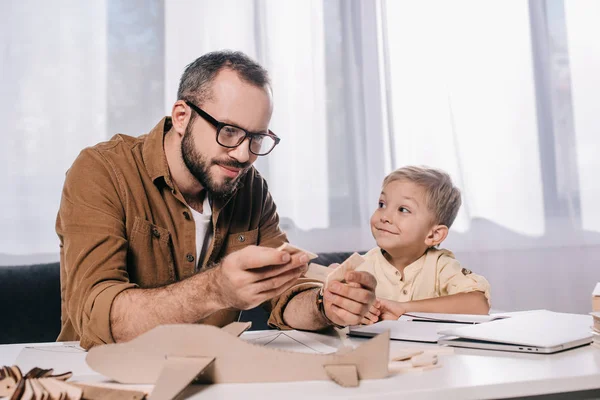 Entzückend glücklicher Sohn schaut lächelnden Vater beim Modellieren von Spielzeugflugzeugen zu Hause an — Stockfoto