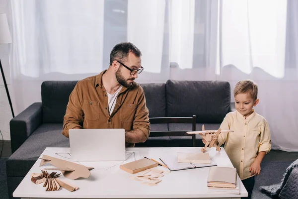 Vater benutzt Laptop und sieht kleinem Sohn zu Hause beim Spielen mit Holzflugzeugmodell zu — Stockfoto