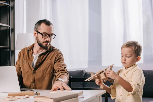 Vater benutzt Laptop, während entzückender kleiner Sohn zu Hause mit hölzernem Flugzeugmodell spielt — Stockfoto