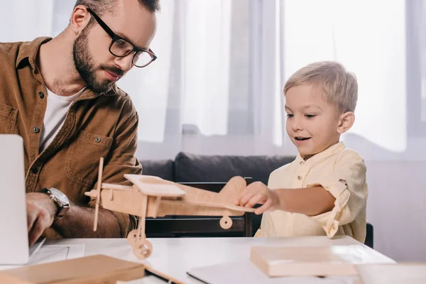 Father using laptop while smiling son playing with wooden plane model — Stock Photo