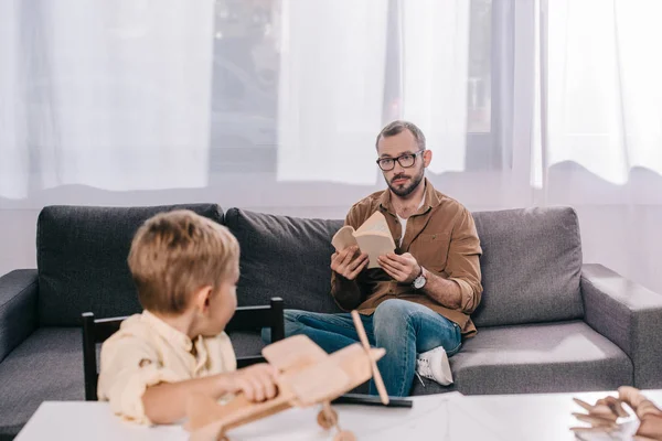 Vater hält Buch in der Hand und schaut kleinem Sohn beim Spielen mit Holzflugzeugmodell zu — Stockfoto