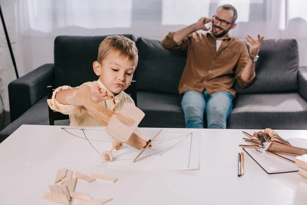 Little son playing with wooden plane model while father talking by smartphone behind — Stock Photo