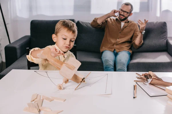 Pequeño hijo jugando con el modelo de avión de madera, mientras que el padre habla por teléfono inteligente detrás - foto de stock