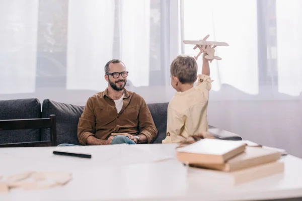 Feliz padre mirando lindo pequeño hijo jugando con juguete de madera avión en casa - foto de stock