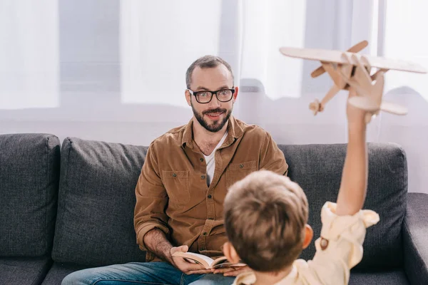 Mignon petit garçon jouer avec jouet avion tout en souriant père lecture livre sur canapé — Photo de stock