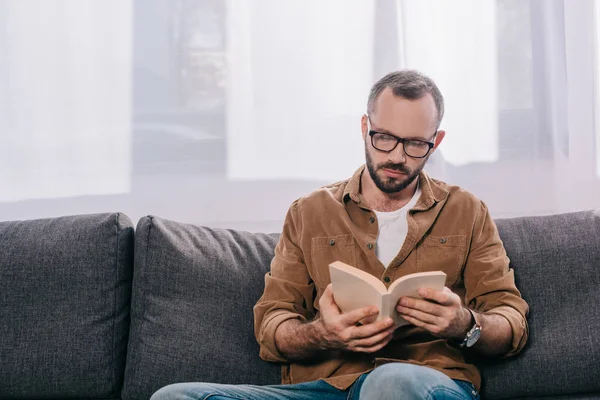 Hombre barbudo guapo en gafas lectura libro en casa - foto de stock