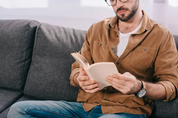 Ritagliato colpo di uomo barbuto in occhiali libro di lettura a casa — Foto stock