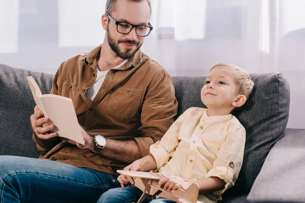 Felice padre tenendo libro e guardando il figlio giocare con aereo giocattolo di legno a casa — Foto stock