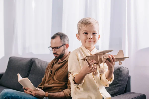 Adorable niño sosteniendo juguete avión de madera y sonriendo a la cámara mientras padre leyendo libro detrás - foto de stock