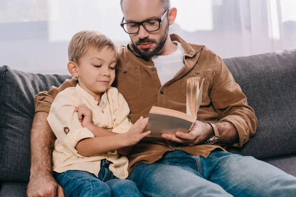 Pai e filho sentados no sofá e lendo livro juntos — Fotografia de Stock
