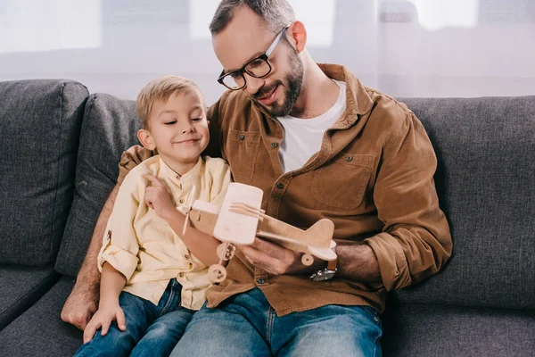 Pai feliz e filho pequeno bonito sentado no sofá e brincando com o modelo de avião de madeira — Fotografia de Stock