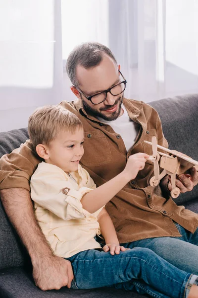 Glücklicher Vater und Sohn sitzen auf der Couch und spielen mit einem hölzernen Flugzeugmodell — Stockfoto