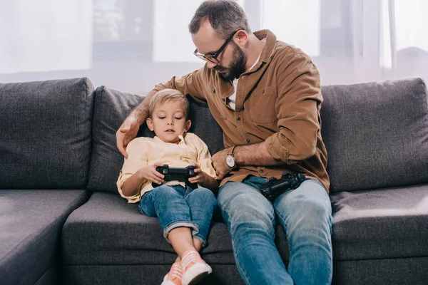 Father looking at cute little son sitting on couch and using joystick — Stock Photo